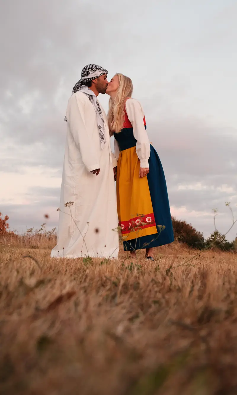 Alice och Yazan kysser varandra i fältet, hon i Varbergsdräkt, han i vit thobe.
